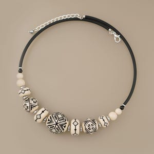 May include: A necklace featuring a black rubber cord with a silver-toned metal lobster clasp and an adjustable chain. The necklace is adorned with cream-colored beads, some of which are decorated with intricate black geometric patterns and designs.