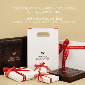 Gift-ready jewelry packaging from Oksana Boriychuk, featuring elegant white boxes tied with red ribbons and optional dark wooden boxes with engraved floral patterns, accompanied by a branded paper gift bag.