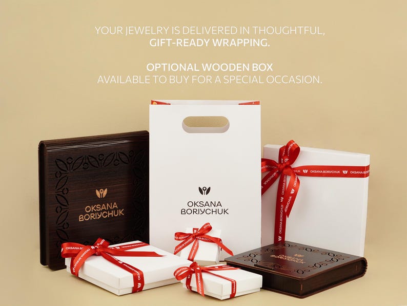 Gift-ready jewelry packaging from Oksana Boriychuk, featuring elegant white boxes tied with red ribbons and optional dark wooden boxes with engraved floral patterns, accompanied by a branded paper gift bag.