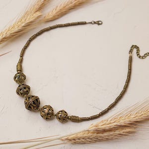 May include: A brass-toned necklace featuring a series of openwork spherical beads interspersed with smaller, cylindrical beads. The necklace is accented with a chain and clasp closure. Wheat stalks are arranged around the necklace on a neutral background.