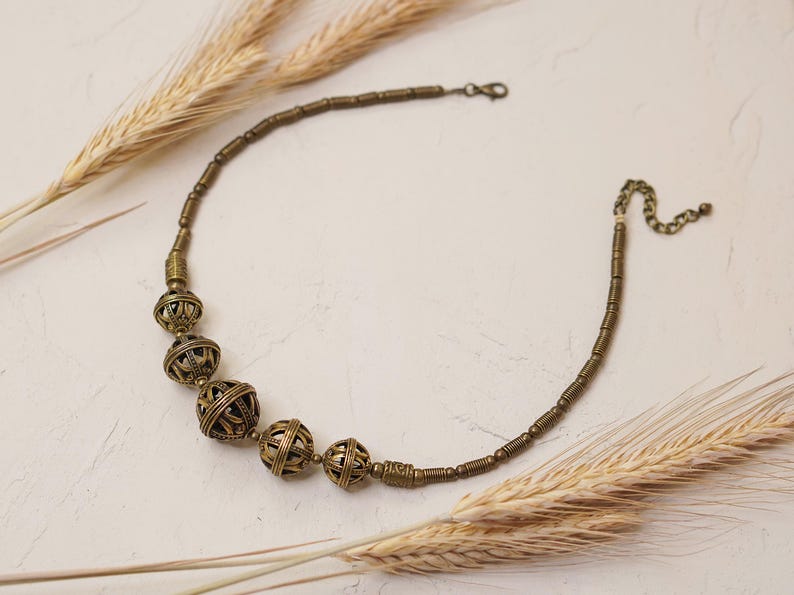 May include: A brass-toned necklace featuring a series of openwork spherical beads interspersed with smaller, cylindrical beads. The necklace is accented with a chain and clasp closure. Wheat stalks are arranged around the necklace on a neutral background.