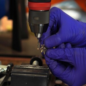 Close-up of a gloved hand drilling a small hole in a star-shaped brass charm using a bench drill, showing the metalworking process in a jewelry workshop.