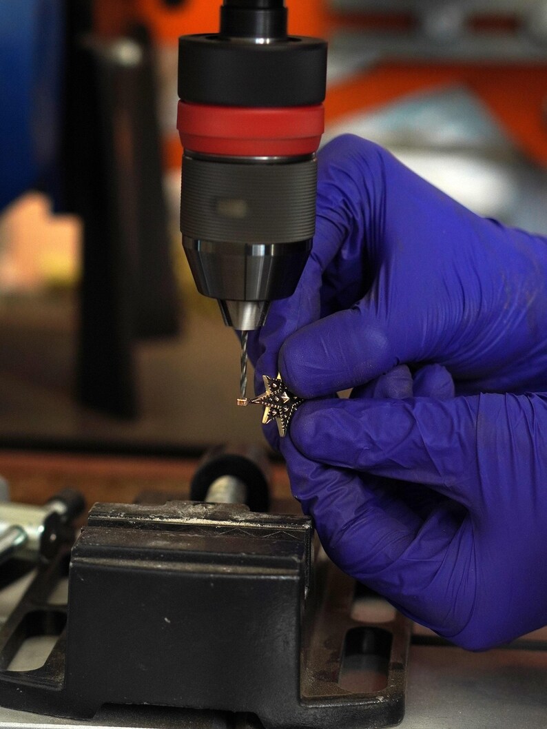 Close-up of a gloved hand drilling a small hole in a star-shaped brass charm using a bench drill, showing the metalworking process in a jewelry workshop.