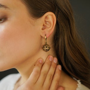 May include: A close-up of a gold-colored earring with a spherical, cage-like design. The earring is suspended from a small hoop. The jewelry is worn on the ear of a person with long brown hair.