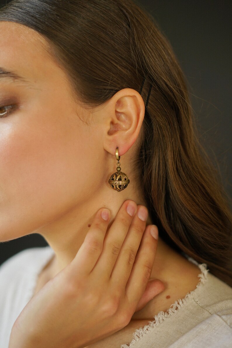 May include: A close-up of a gold-colored earring with a spherical, cage-like design. The earring is suspended from a small hoop. The jewelry is worn on the ear of a person with long brown hair.
