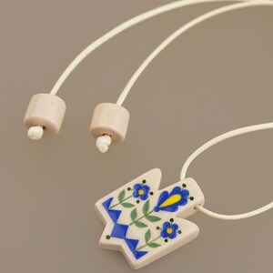 Unique Ukrainian Tryzub pendant necklace featuring a folk art floral design where blue is the dominant color, hand-painted on a cream ceramic base with an adjustable wrap cord.