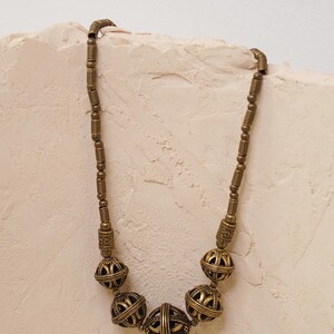 May include: A vintage-style necklace with a brass finish. The necklace features a series of round, openwork beads interspersed with smaller, cylindrical beads. The beads have an aged appearance, suggesting a handcrafted or antique origin. The necklace is displayed against a neutral background.
