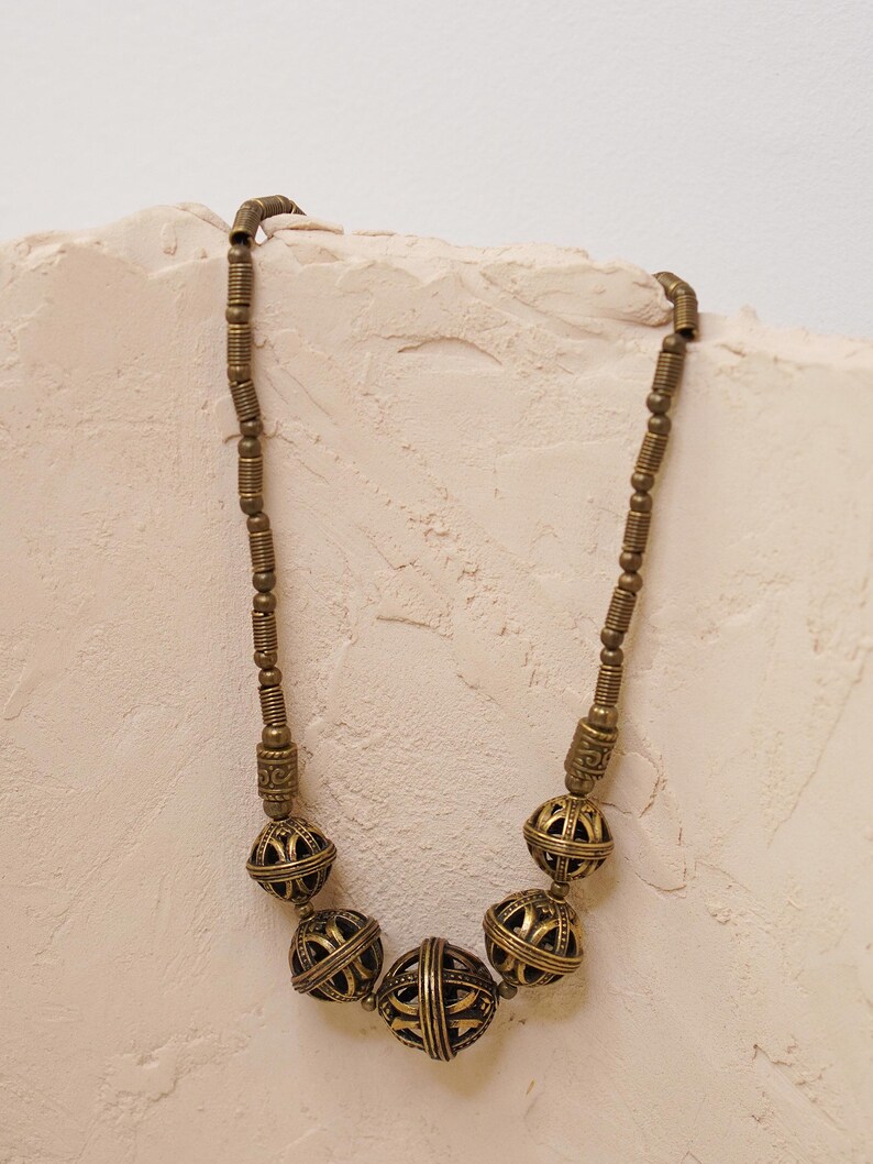 May include: A vintage-style necklace with a brass finish. The necklace features a series of round, openwork beads interspersed with smaller, cylindrical beads. The beads have an aged appearance, suggesting a handcrafted or antique origin. The necklace is displayed against a neutral background.