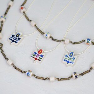 Assortment of ceramic Tryzub pendants in different folk floral color variations—navy-dominant, red-dominant, and light blue—displayed with matching beaded necklaces and cream cord options.