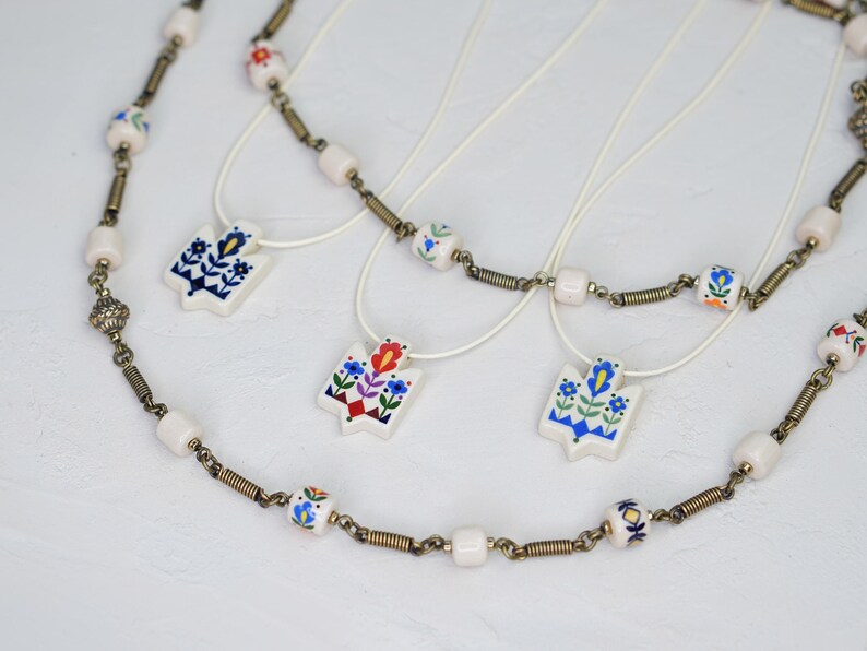 Assortment of ceramic Tryzub pendants in different folk floral color variations—navy-dominant, red-dominant, and light blue—displayed with matching beaded necklaces and cream cord options.