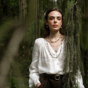 Person standing in a forest wearing layered folk-inspired necklaces, including a ceramic Tryzub pendant with a blue floral design, styled with a white blouse and dark trousers.