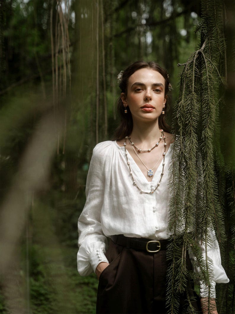 Person standing in a forest wearing layered folk-inspired necklaces, including a ceramic Tryzub pendant with a blue floral design, styled with a white blouse and dark trousers.