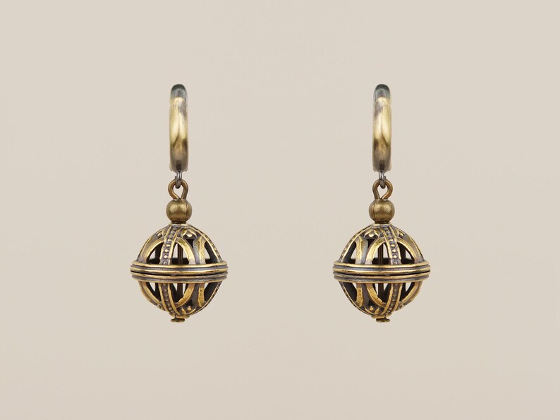 May include: A pair of antique gold-colored earrings. Each earring features a hoop, a small ball, and a larger, openwork sphere with intricate geometric patterns. The earrings are suspended against a neutral background.