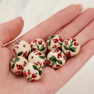 May include: A collection of eight round, cream-coloured beads featuring a hand-painted pattern of small red berries and green leaves. Each bead has a central hole for threading, and the surface displays a smooth, glossy finish with consistent botanical motifs.