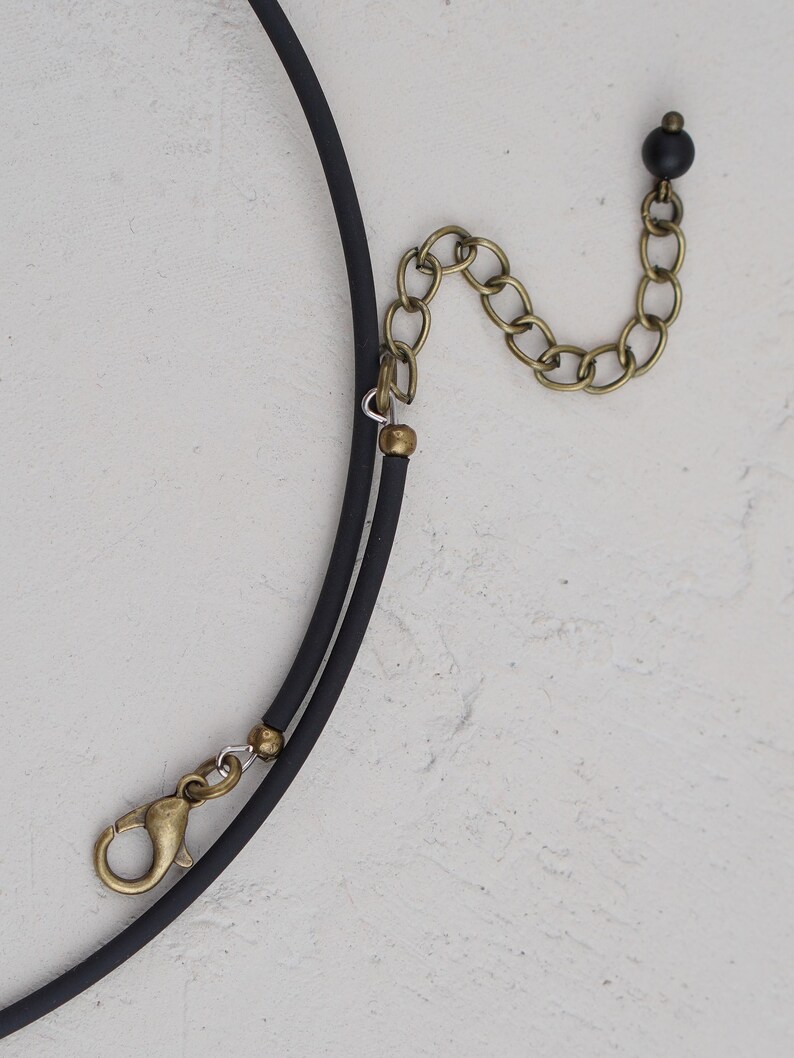 May include: A black choker necklace with a gold chain extender and a small black bead. The necklace is made of a soft, flexible material.