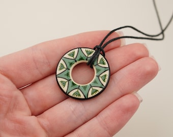 May include: A circular pendant necklace with a black cord. The pendant has a cream-coloured centre and a black border. The design features green and cream-coloured geometric shapes. The necklace is held in a hand.