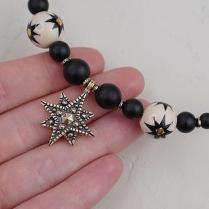 May include: A black and white star-shaped pendant hangs from a necklace made of black and white beads. The pendant is made of metal and has a detailed design.