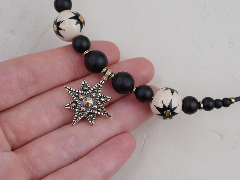 May include: A black and white star-shaped pendant hangs from a necklace made of black and white beads. The pendant is made of metal and has a detailed design.