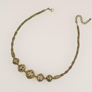 May include: A gold-toned necklace with a variety of beads. The necklace features several large, openwork spherical beads interspersed with smaller, cylindrical beads. The clasp and chain are also gold-toned. The necklace is displayed against a white background.