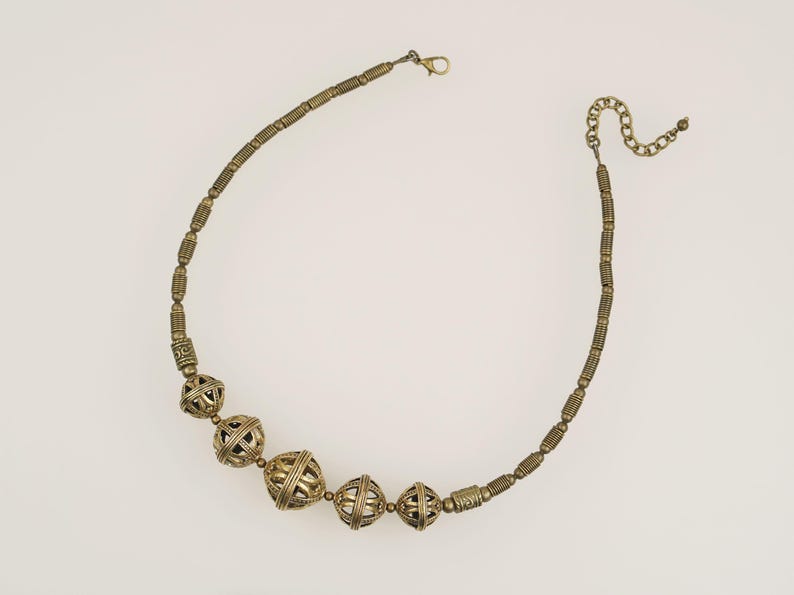 May include: A gold-toned necklace with a variety of beads. The necklace features several large, openwork spherical beads interspersed with smaller, cylindrical beads. The clasp and chain are also gold-toned. The necklace is displayed against a white background.