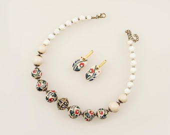 May include: A necklace and earring set featuring white beads, cream-colored spheres, and round beads painted with red flowers and green leaves. The necklace includes a central bronze-toned metal cage bead and a lobster clasp closure.