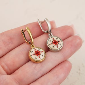 May include: Two round dangle earrings with a cream backdrop and a red starburst design. One earring has a gold-coloured frame and clasp, the other a silver-coloured frame and clasp. The earrings are displayed on a light background.