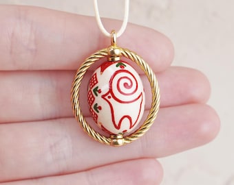 May include: A gold pendant necklace with a white and red egg-shaped charm. The charm has a red and white swirl design and a small green floral pattern. The charm is suspended in a gold ring.