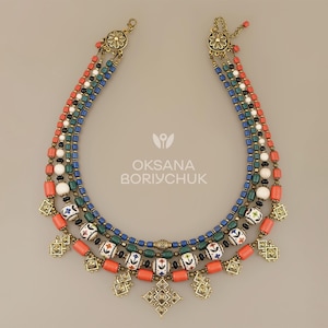 May include: A layered necklace with coral, white, and blue beads, and gold-coloured metal details. The necklace has a floral pattern and the text "OKSANA BORYCHUK".