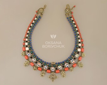 May include: A multi-strand necklace featuring coral, white, and blue beads, accented with gold-toned metalwork. The necklace has a floral design and the text "OKSANA BORYCHUK".