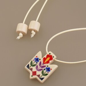 Unique Ukrainian Tryzub pendant necklace featuring a folk art floral design where red is the dominant color, hand-painted on a cream ceramic base with an adjustable wrap cord.