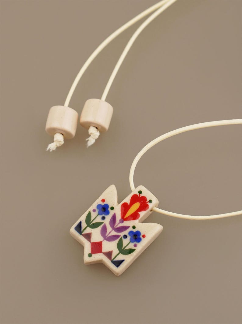Unique Ukrainian Tryzub pendant necklace featuring a folk art floral design where red is the dominant color, hand-painted on a cream ceramic base with an adjustable wrap cord.