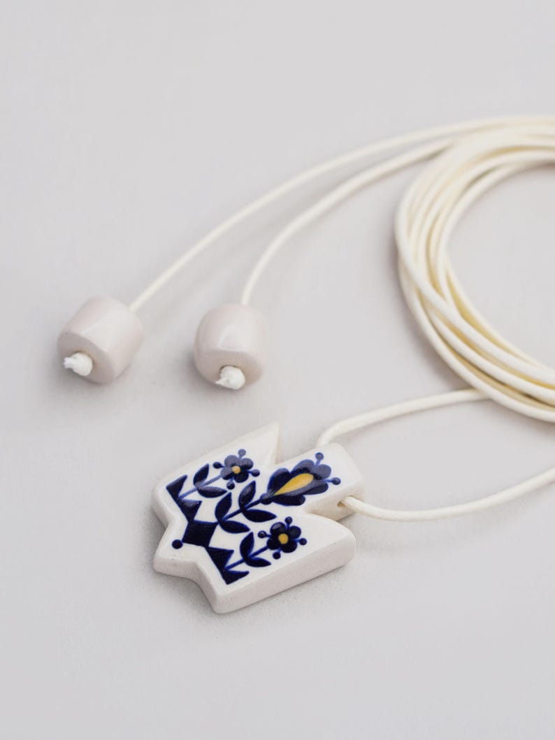 Handmade ceramic Tryzub pendant necklace with floral folk art design in navy blue and yellow on a white base, featuring an adjustable cream cord with ceramic bead ends.