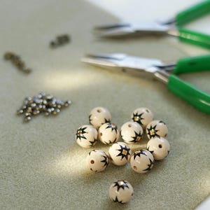 Hand-painted ceramic beads with an earthy white glaze and black star folk motifs laid out on a work surface, with small metal findings and green-handled jewelry pliers visible in the background, showing the handmade jewelry-making process.