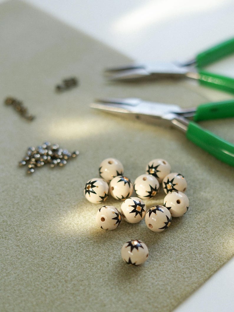 Hand-painted ceramic beads with an earthy white glaze and black star folk motifs laid out on a work surface, with small metal findings and green-handled jewelry pliers visible in the background, showing the handmade jewelry-making process.