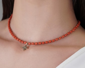 May include: A coral-colored beaded necklace with a gold-toned pendant. The pendant features a geometric design with black accents. The necklace is worn around the neck, showcasing the jewelry's design and color.