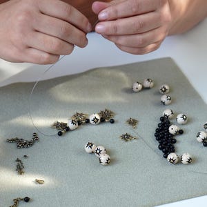 Hands assembling a handmade necklace by stringing beads on wire, with hand-painted ceramic beads featuring black star motifs, small black beads, brass star charms, and metal findings arranged on a soft work mat in a jewelry studio.