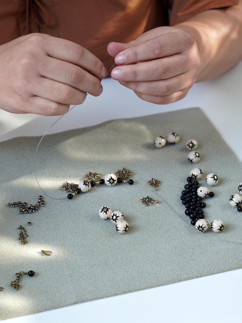 Hands assembling a handmade necklace by stringing beads on wire, with hand-painted ceramic beads featuring black star motifs, small black beads, brass star charms, and metal findings arranged on a soft work mat in a jewelry studio.