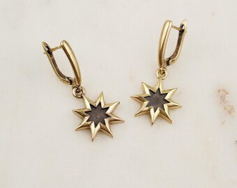 May include: A pair of gold-colored starburst earrings. Each earring features a star-shaped pendant with a dark center and a gold-colored frame. The earrings have a lever-back closure and are displayed on a white marble surface.