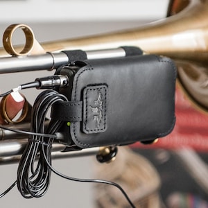 Puede incluir: Una funda de cuero negro con un logotipo está sujeta a un instrumento de latón. La funda tiene una correa y contiene un pequeño dispositivo con cables negros y un conector. La campana del instrumento es visible en el fondo.
