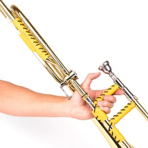 May include: A gleaming brass trombone featuring yellow leather grips with black stitching. The instrument is set against a white backdrop, emphasizing its polished surface and detailed construction. The trombone's slide and bell are visible, highlighting its musical components.