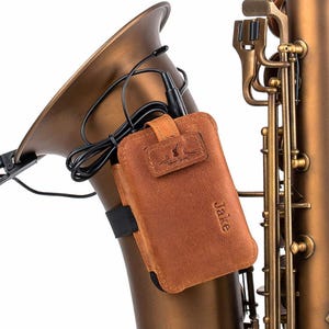 May include: A bronze saxophone with a brown leather case secured to it. The case is embossed with the name "Jake" and a logo. A black cable and microphone are connected to the instrument, highlighting its details.