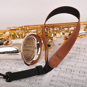 May include: A gold and silver saxophone with a brown leather neck strap. The saxophone rests on a sheet of musical notes. The instrument has gold keys and a silver body.