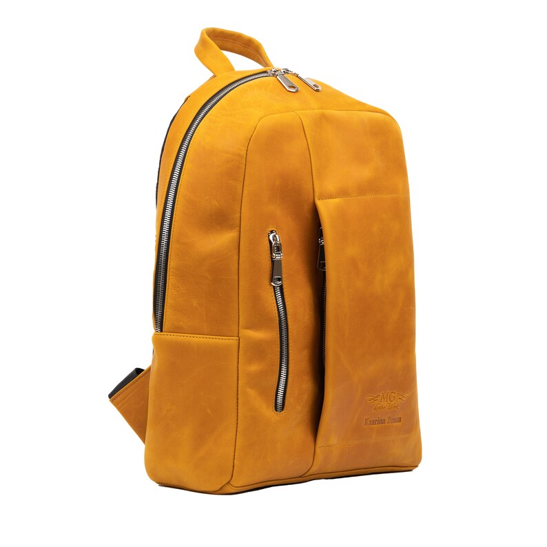Puede incluir: Una mochila de cuero amarillo mostaza con un bolsillo con cremallera en la parte delantera y un tirador de cremallera plateado. La mochila tiene un peque&ntilde;o bolsillo en el lateral y el nombre de la marca "MG" est&aacute; impreso en la parte delantera.
