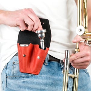 May include: A red leather pouch with two compartments for holding trombone mouthpieces. A silver mouthpiece is partially inserted into the pouch. A person is holding a trombone in the background.