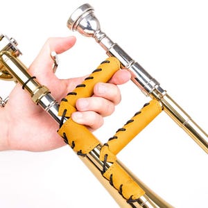 Op de afbeelding: Een koperen trombone met een zilveren mondstuk en een gele leren grip met zwarte stiksels. Het instrument wordt door een hand tegen een witte achtergrond gehouden.