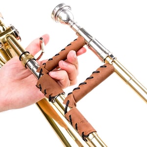 May include: A brass trombone with a brown leather grip stitched with black thread. The trombone is partially visible, with the slide and bell section in view. The leather grip is designed to enhance the player's hold and comfort.