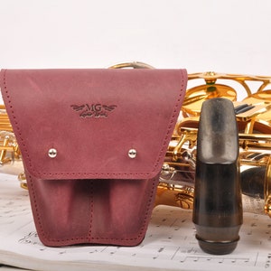 Peut inclure: Un étui à anche de saxophone en cuir bordeaux avec un design cousu et un petit logo qui dit "MG Good Music". L'étui est posé sur une feuille de musique avec un saxophone doré et un bec noir.