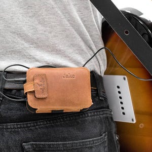 May include: A brown leather case with the name "Jake" embossed on the front, attached to a belt. The case has a flap with a decorative design and is connected to a guitar. The case is approximately 4 inches wide.