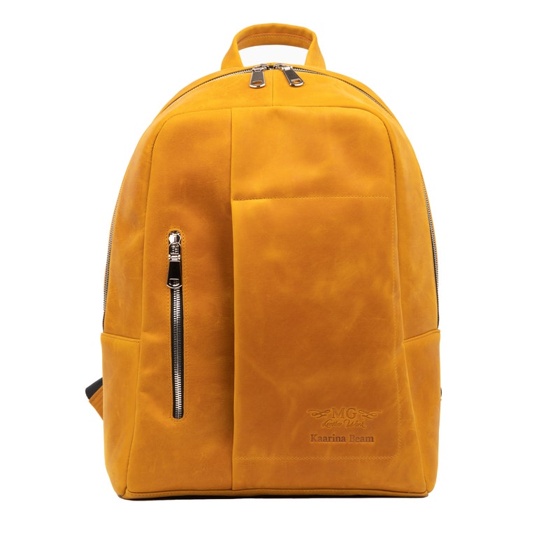 Puede incluir: Una mochila de cuero amarillo mostaza con un bolsillo frontal grande y un bolsillo lateral con cremallera. La mochila tiene un peque&ntilde;o logotipo que dice "MG Leather" y "Karina Beam".