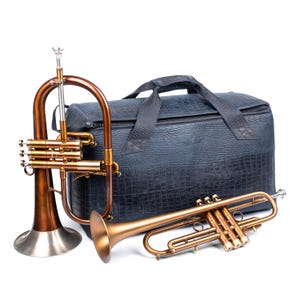 May include: A flugelhorn and a trumpet in a warm bronze finish are displayed next to a dark blue, textured carrying case. The flugelhorn has a silver bell and the trumpet is a smaller size. Musical instruments.